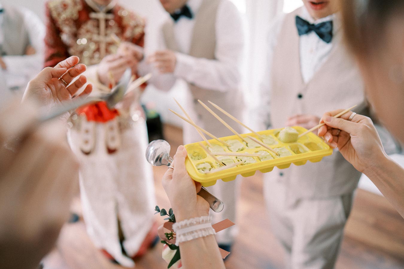 Group of people participating in a wedding game holding skewers with wasabi balls in an ice tray, wearing formal attire.
