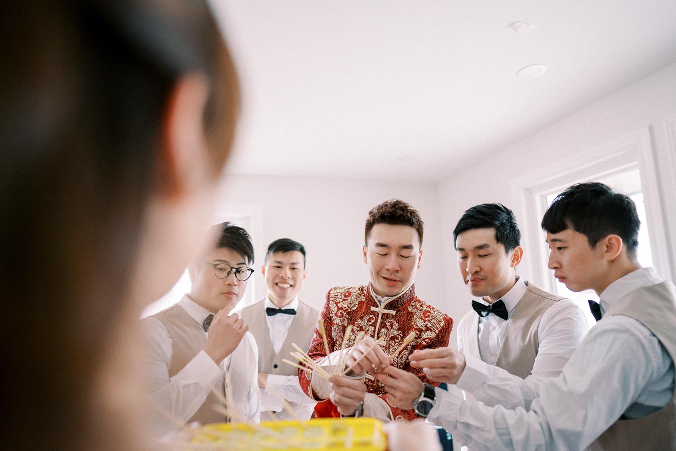 A group of groomsmen in formal attire playing a fun wedding game indoors, with one wearing an intricately designed traditional Chinese jacket, highlighting pre-ceremony traditions.