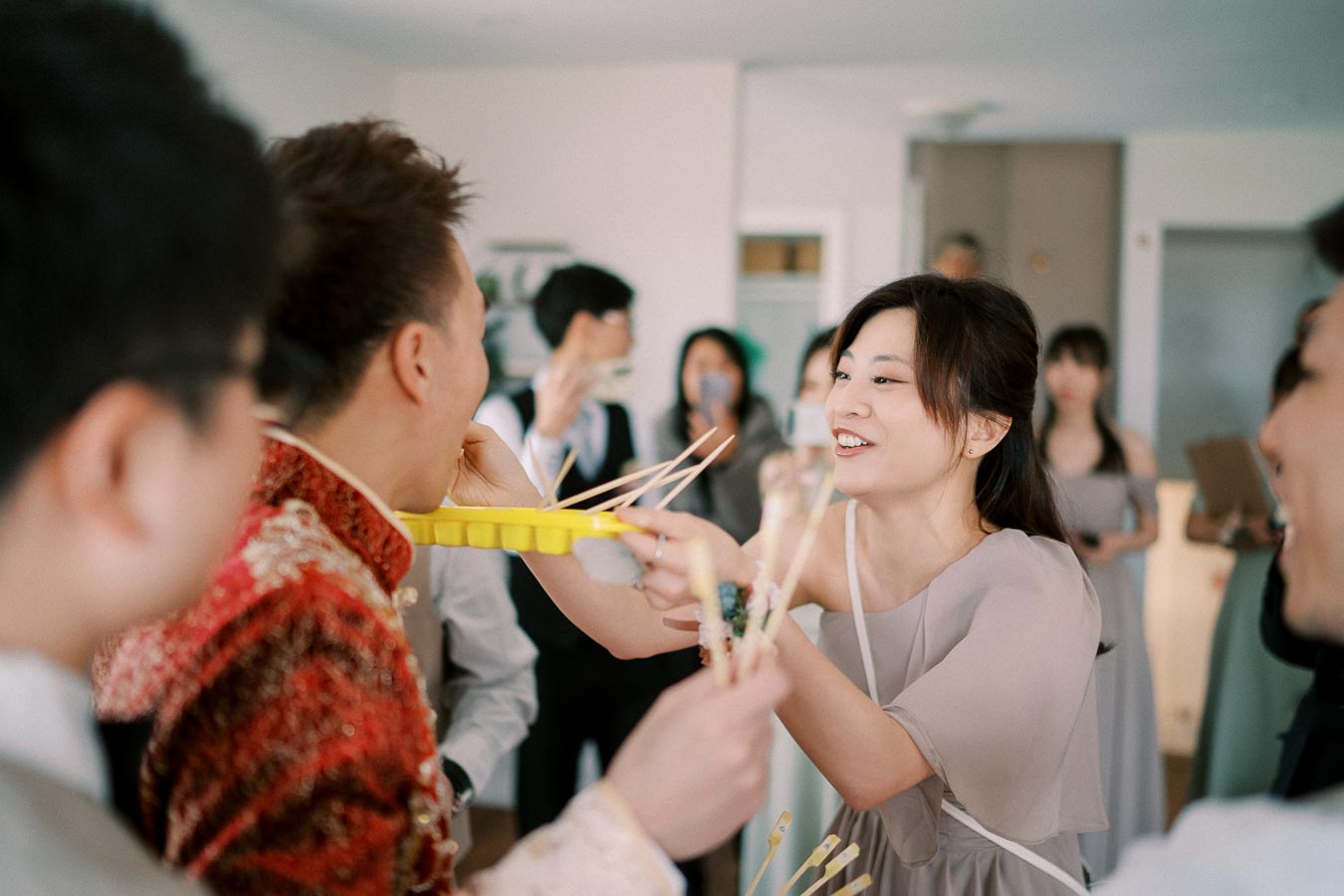 A woman in a gray dress playfully feeds a man with chopsticks during a lively gathering, surrounded by smiling guests in a bright room.
