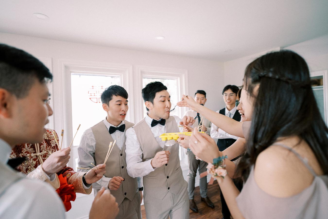 A group of groomsmen in beige suits play a fun wedding game indoors, using chopsticks to pick up small items while a bridesmaid assists, creating a joyful and lively atmosphere.