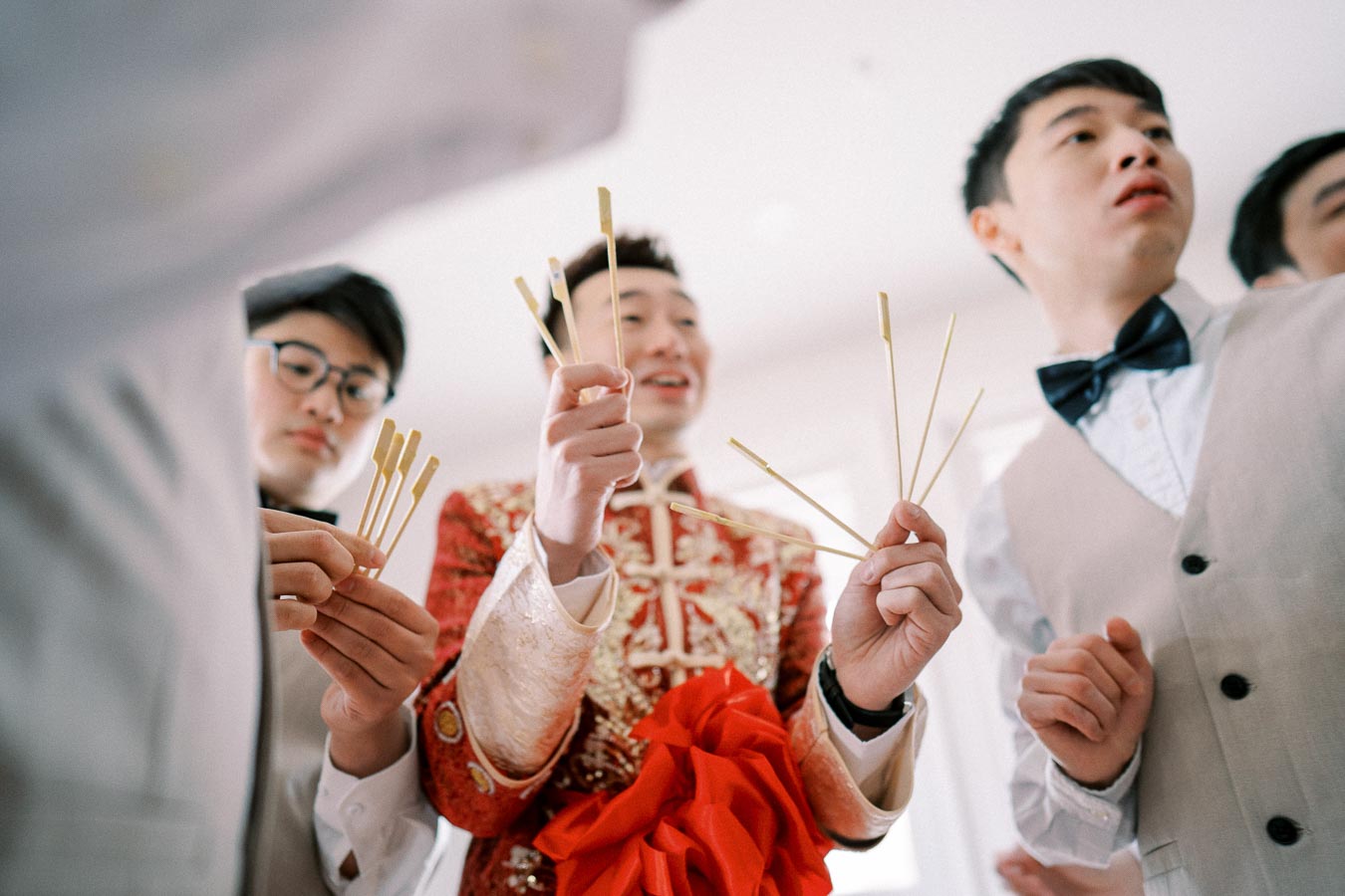 A group of men dressed in formal attire holding traditional Chinese wedding sticks, with one wearing an ornate red and gold outfit, suggesting a cultural wedding ceremony.