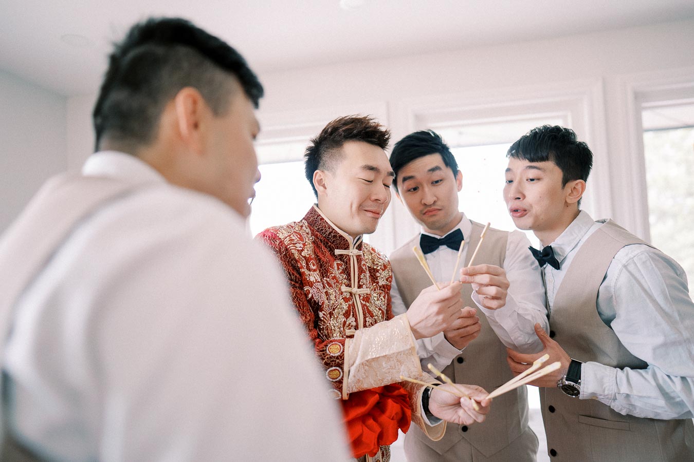 A group of young men engaged in a playful activity, with one wearing a traditional red embroidered outfit and others in beige vests, holding chopsticks in a well-lit room.