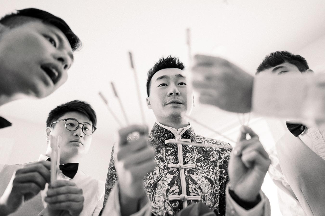 Group of men in formal attire participating in a traditional ceremony, holding sticks, with the focus on a man in an ornate embroidered outfit. Black and white image captures cultural celebration.