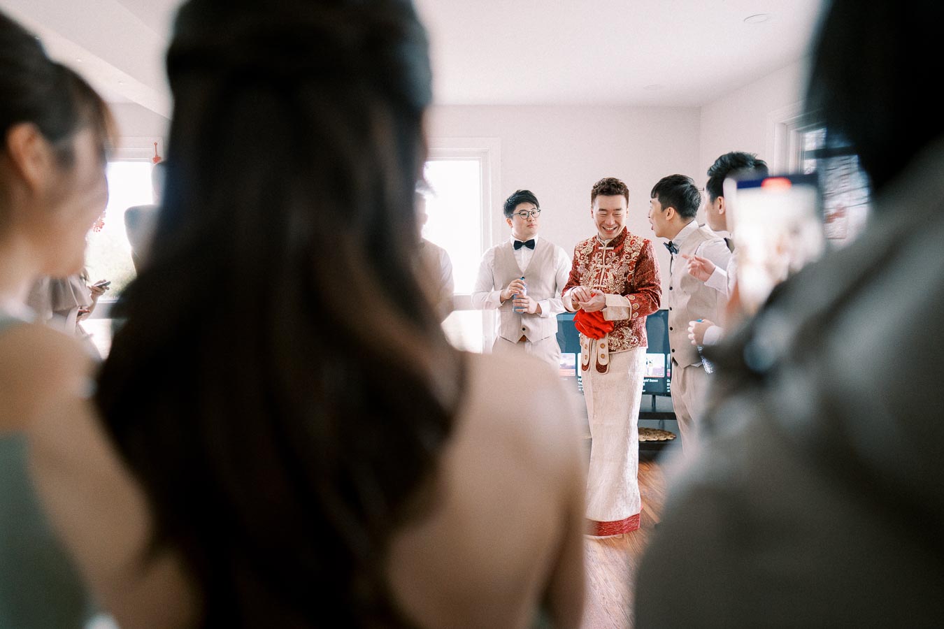 A joyful groom in traditional red attire is surrounded by groomsmen in light suits during a wedding preparation in a bright room.