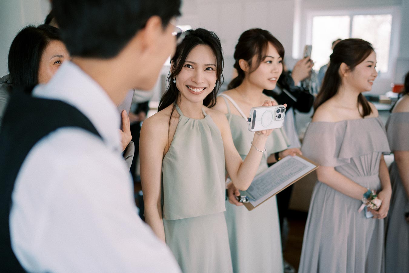 A group of bridesmaids in elegant dresses smiling and capturing a moment with a smartphone at a wedding event.