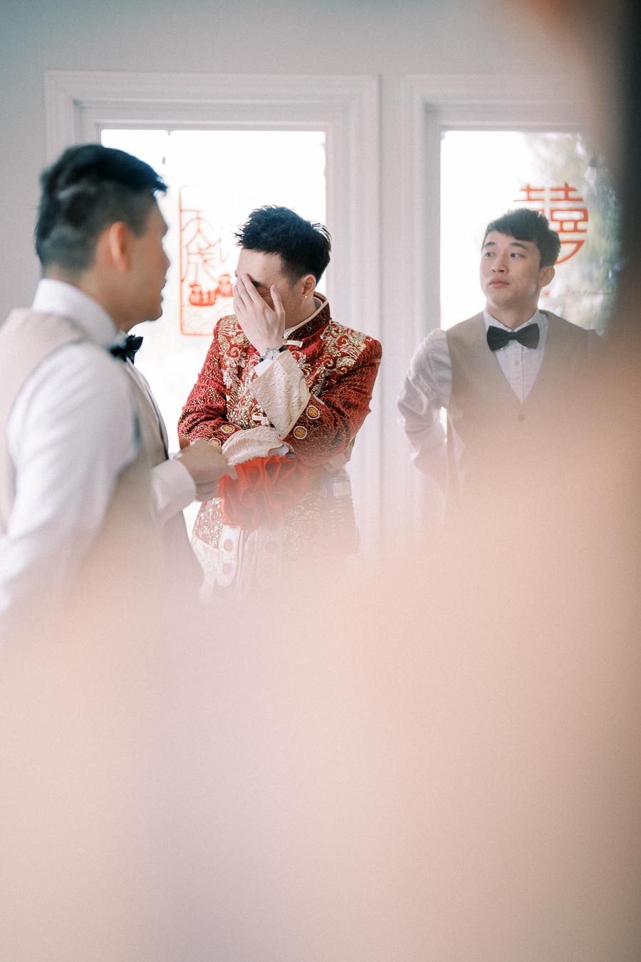 A groom in traditional red attire covers his face with his hand, standing between two groomsmen in formal wear, inside a room with decorations on the glass doors.