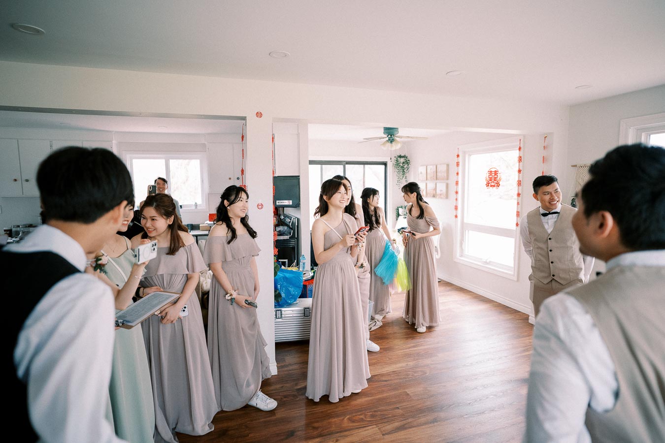 Group of bridesmaids in taupe dresses and groomsmen in beige suits sharing a joyful moment in a bright, decorated living room.