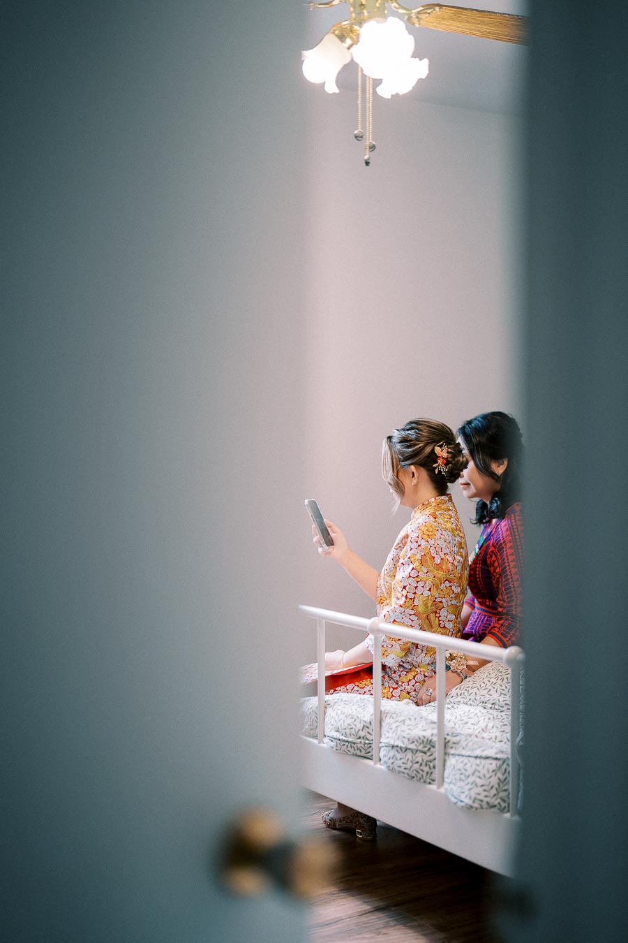 Two women in traditional clothing sitting on a bed, one holding a smartphone, viewed through a partially open door.