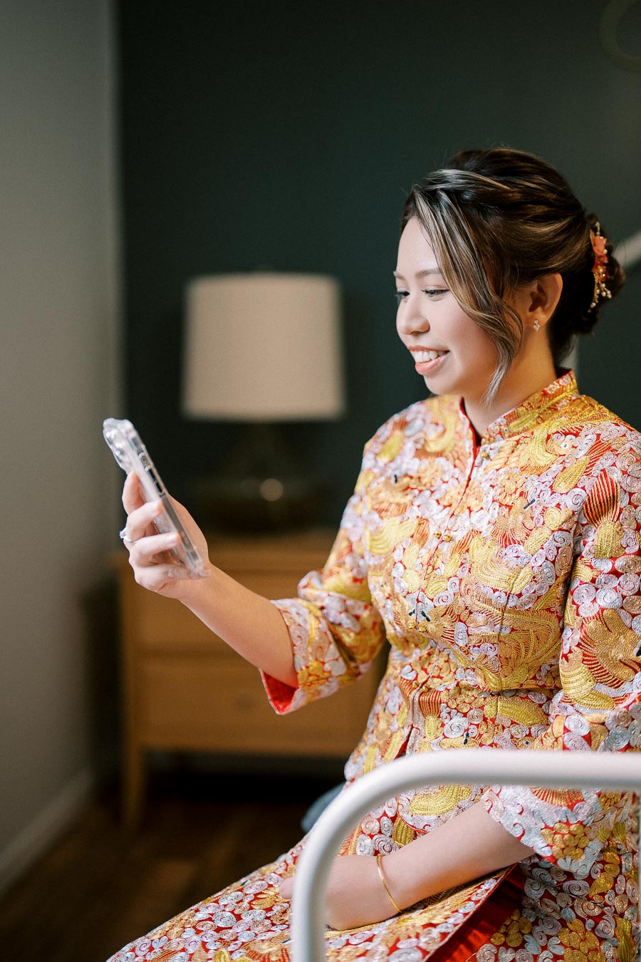 A woman in an embroidered traditional dress smiling at her smartphone indoors.