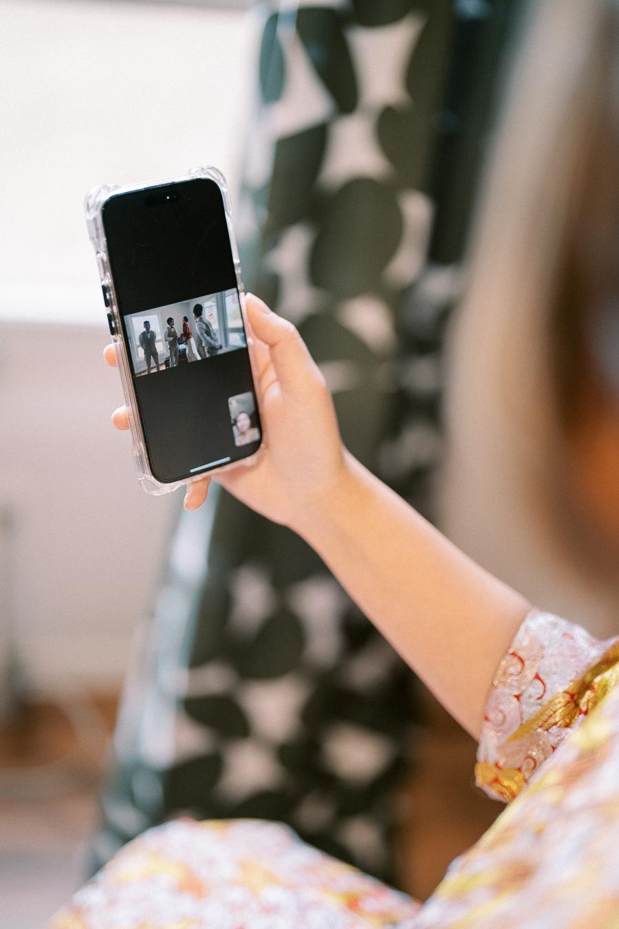 A person holding a smartphone displaying a video call with multiple participants, in a cozy indoor setting.