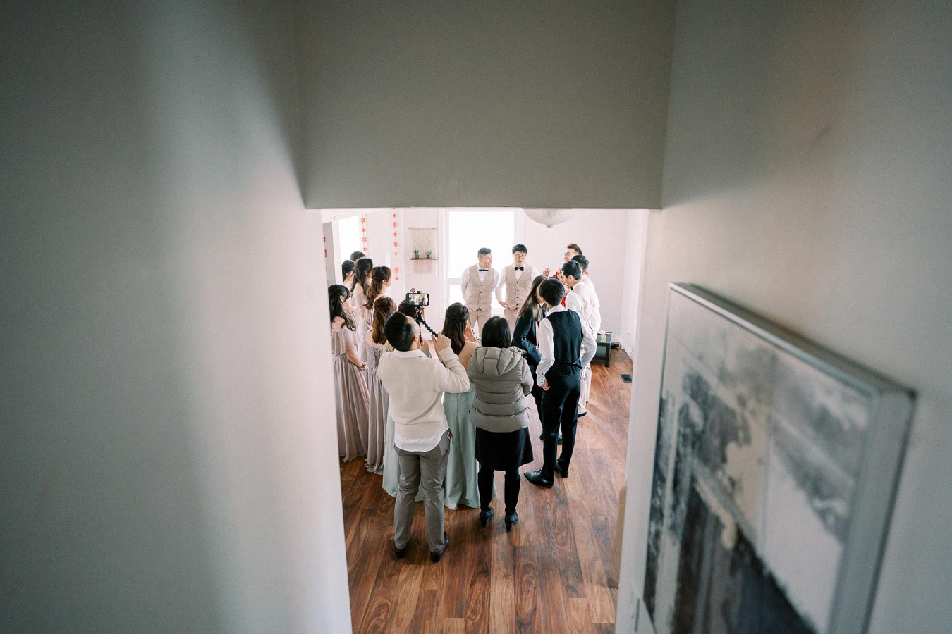 Group of people gathered in a brightly lit room, some dressed formally, possibly preparing for an event or ceremony. The scene is captured from a hallway with a view of wooden flooring, highlighting a sense of anticipation and organization.