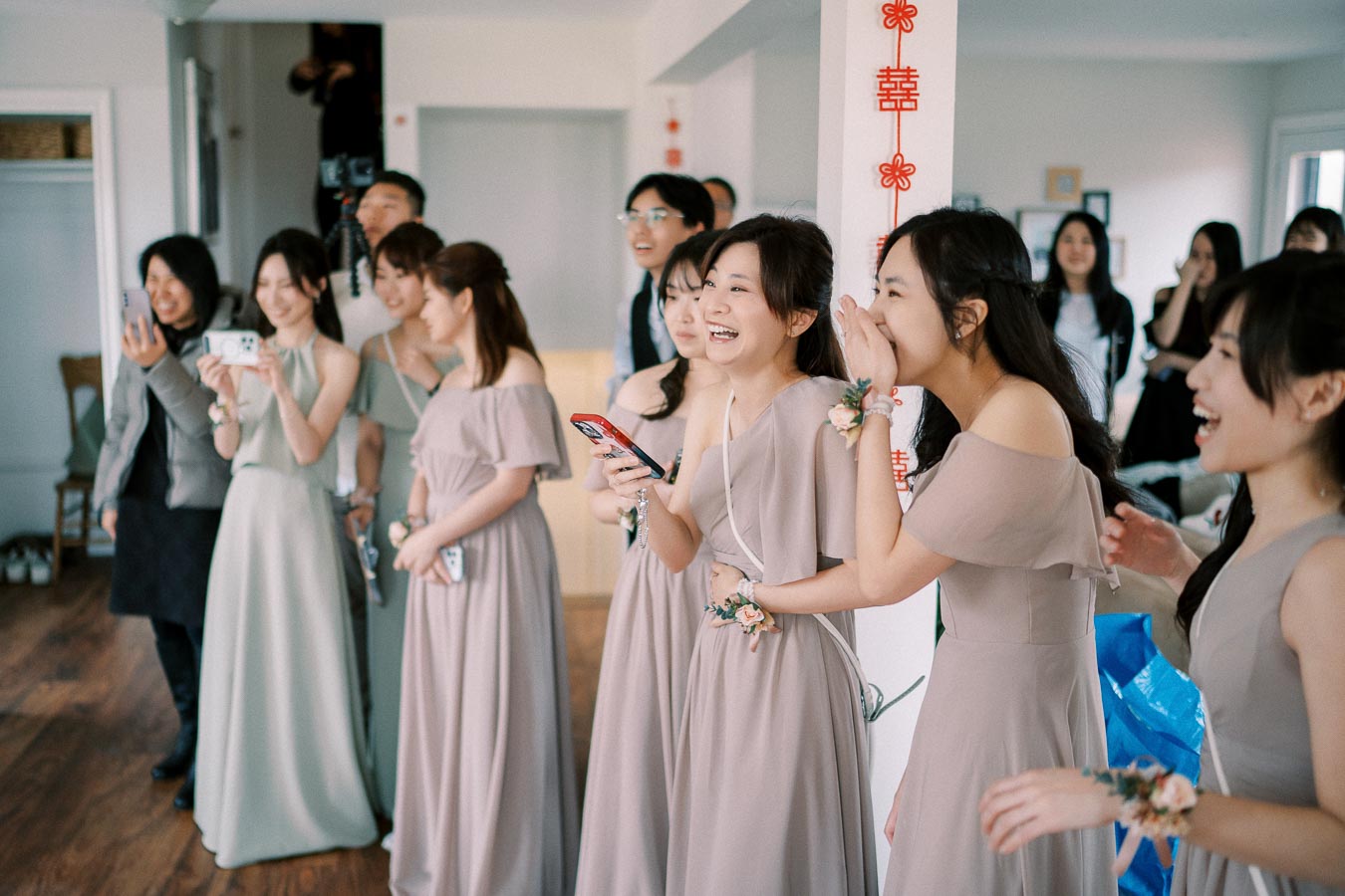 A group of bridesmaids in matching dresses laughing and taking photos during a lively moment at a wedding celebration inside a decorated room.