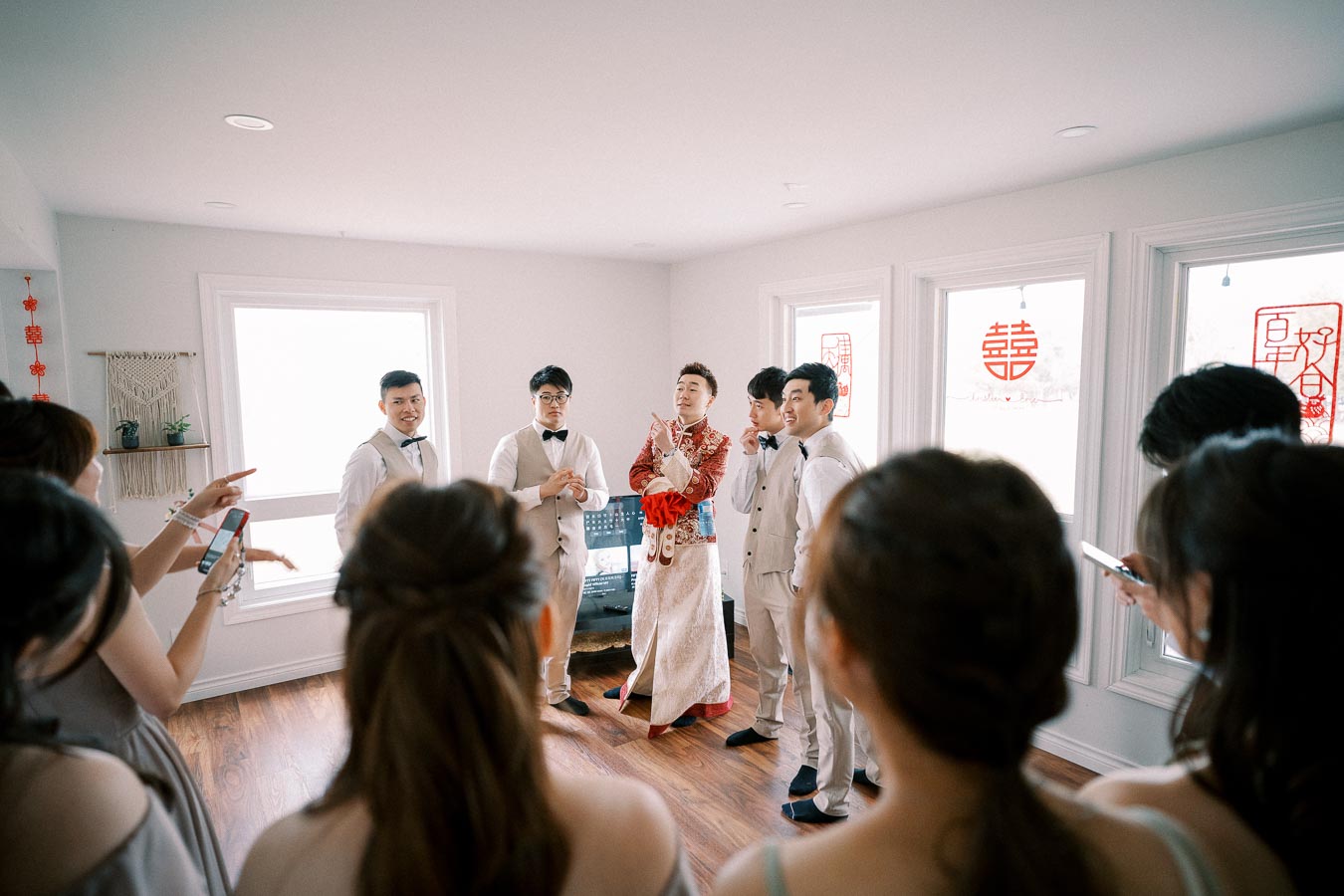 Group of people participating in a traditional wedding ceremony indoors, with the groom in ceremonial attire and groomsmen in matching suits, surrounded by bridesmaids and festive decorations.