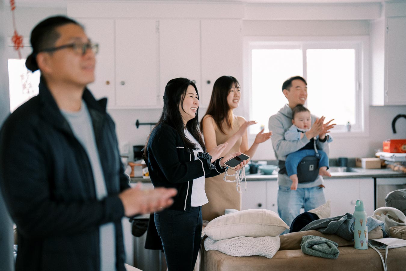 A group of four people, including a man holding a baby, stand in a bright kitchen clapping and smiling, suggesting a joyful gathering or celebration.