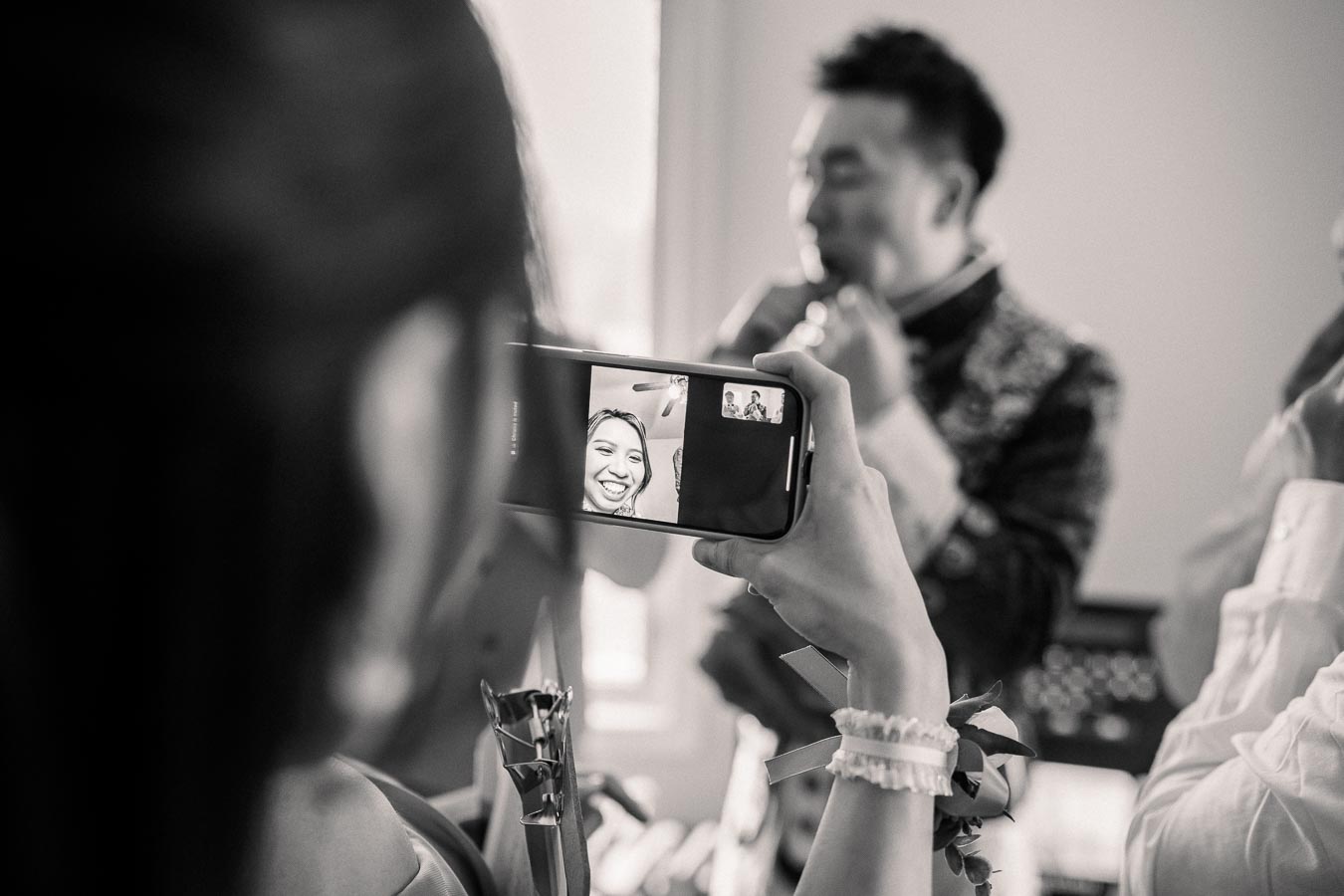 Black and white image of a person holding a smartphone capturing a video call, with a smiling person on the screen, and a blurred background of others getting ready, possibly for a celebration or event.