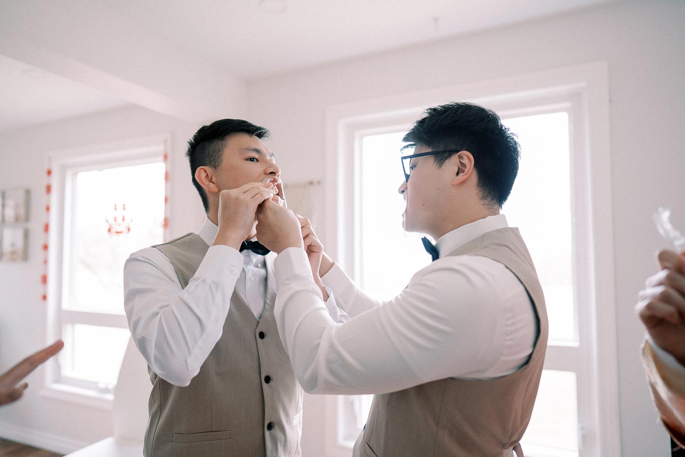 Two men in formal attire, one assisting the other with his bow tie, in a bright room with large windows.
