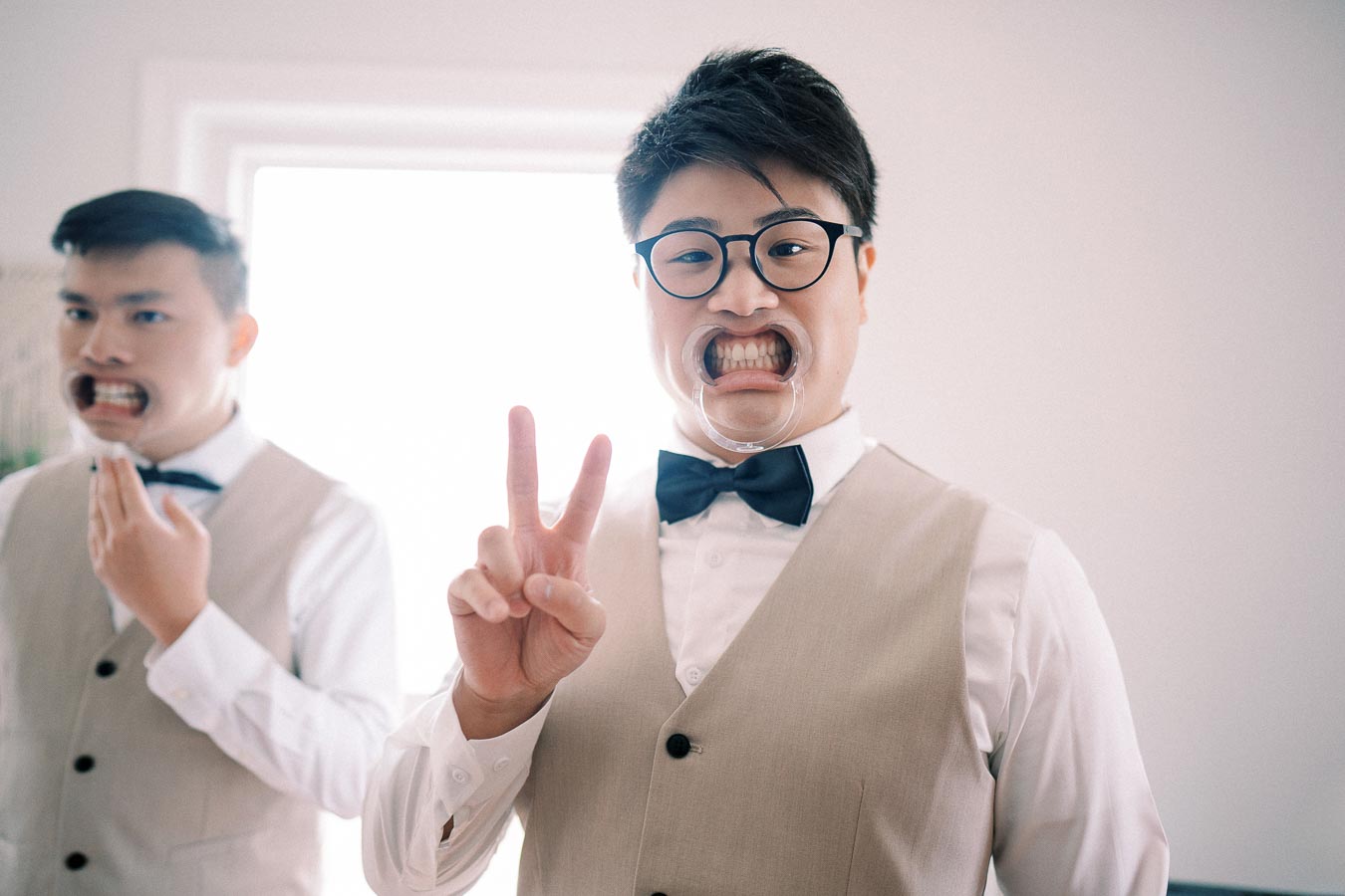Two men in formal attire wearing mouth stretchers, with one making a peace sign.