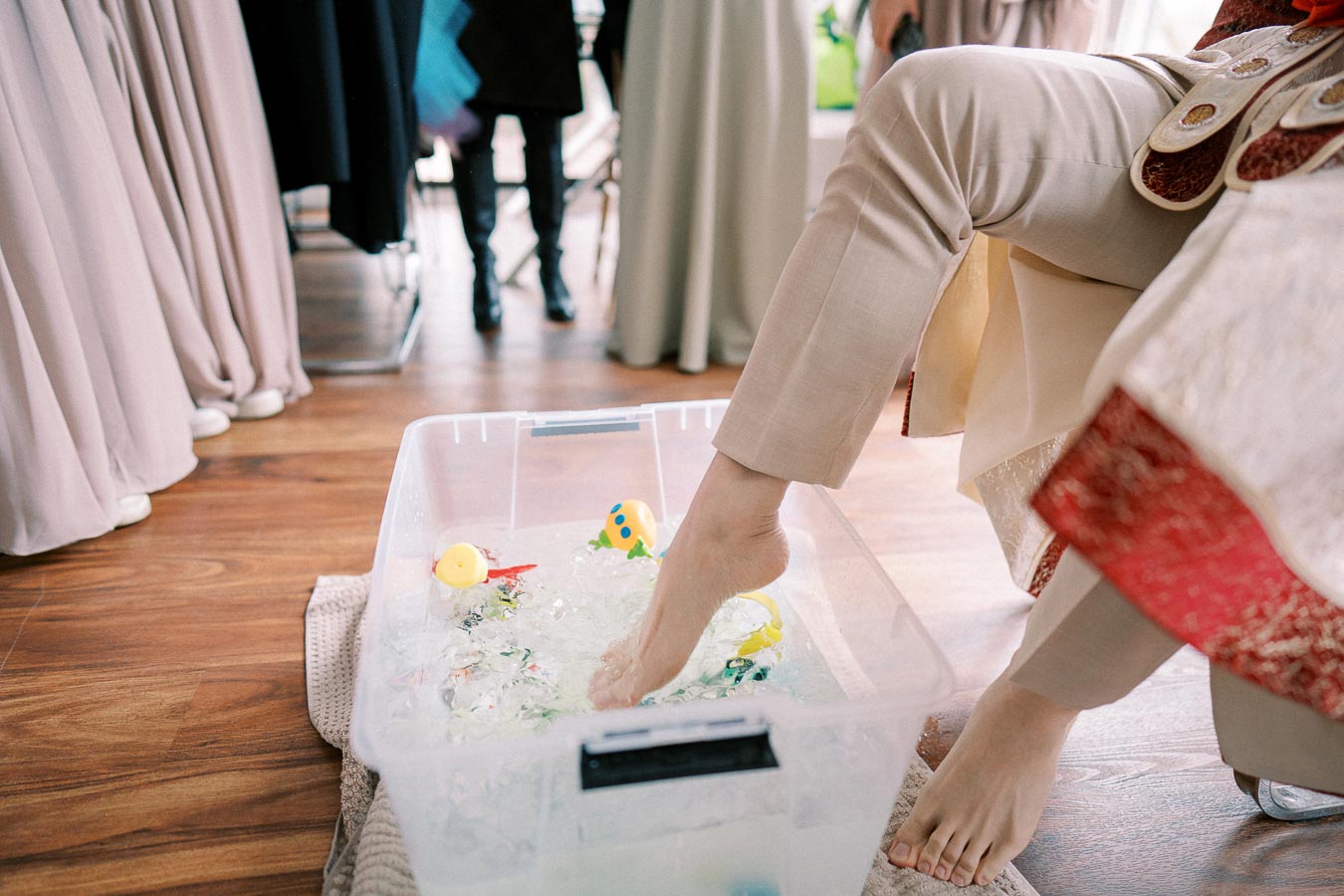 Person soaking foot in a plastic tub filled with ice and rubber ducks, wearing beige pants and embroidered garments, on a wooden floor with people in the background.