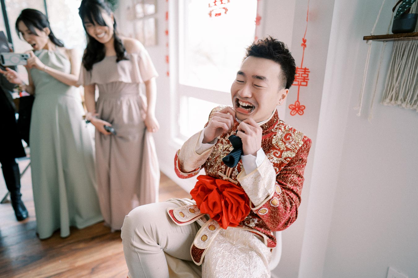 A groom in a traditional Chinese wedding outfit laughs while participating in a fun wedding game. Two bridesmaids, dressed in pastel dresses, smile and watch the moment, enhancing the festive atmosphere. Bright room with Chinese character decorations.