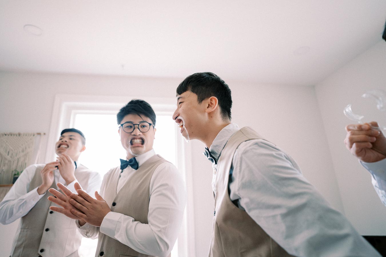 Three men in formal attire laugh while wearing cheek retractors, adding a playful atmosphere to a pre-event gathering. The room is bright with natural light, highlighting their light-colored vests and bow ties.