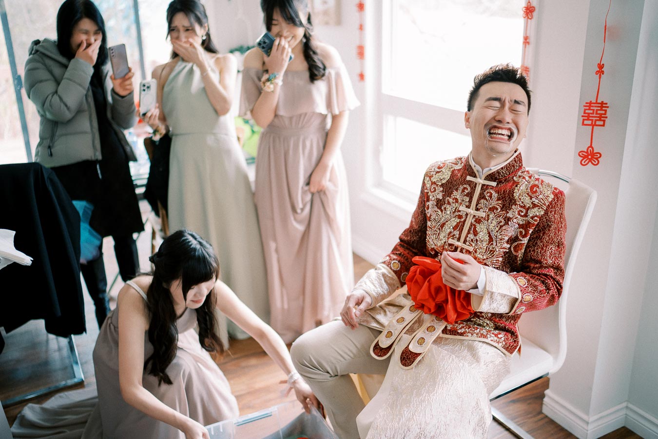 A groom in an ornate red and gold outfit laughs during a Chinese wedding tea ceremony as bridesmaids capture the moment with their phones.