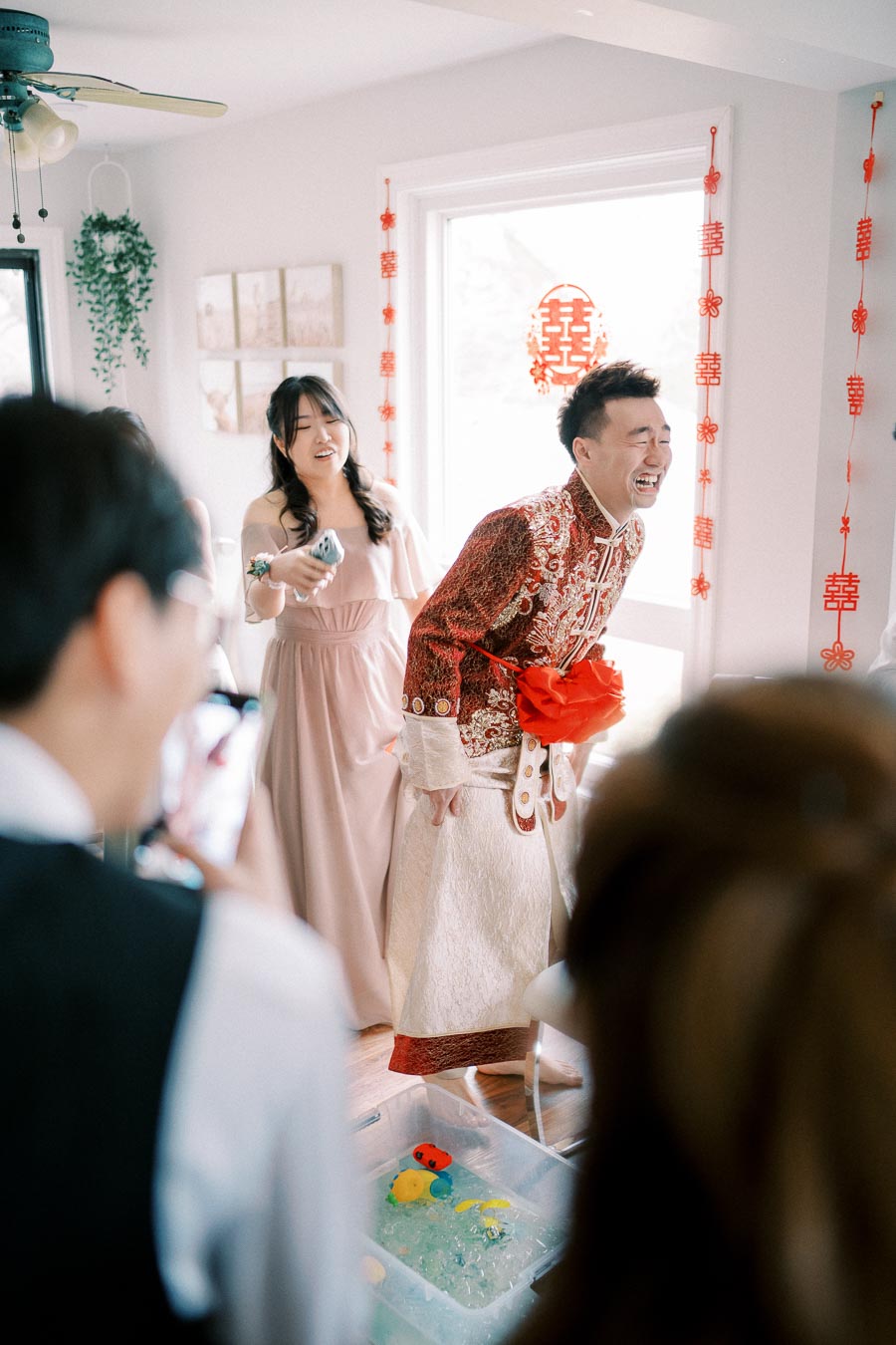 A joyful bride in a pink dress and groom in a traditional embroidered outfit laugh during a playful wedding game, surrounded by festive red decorations and friends capturing the moment.