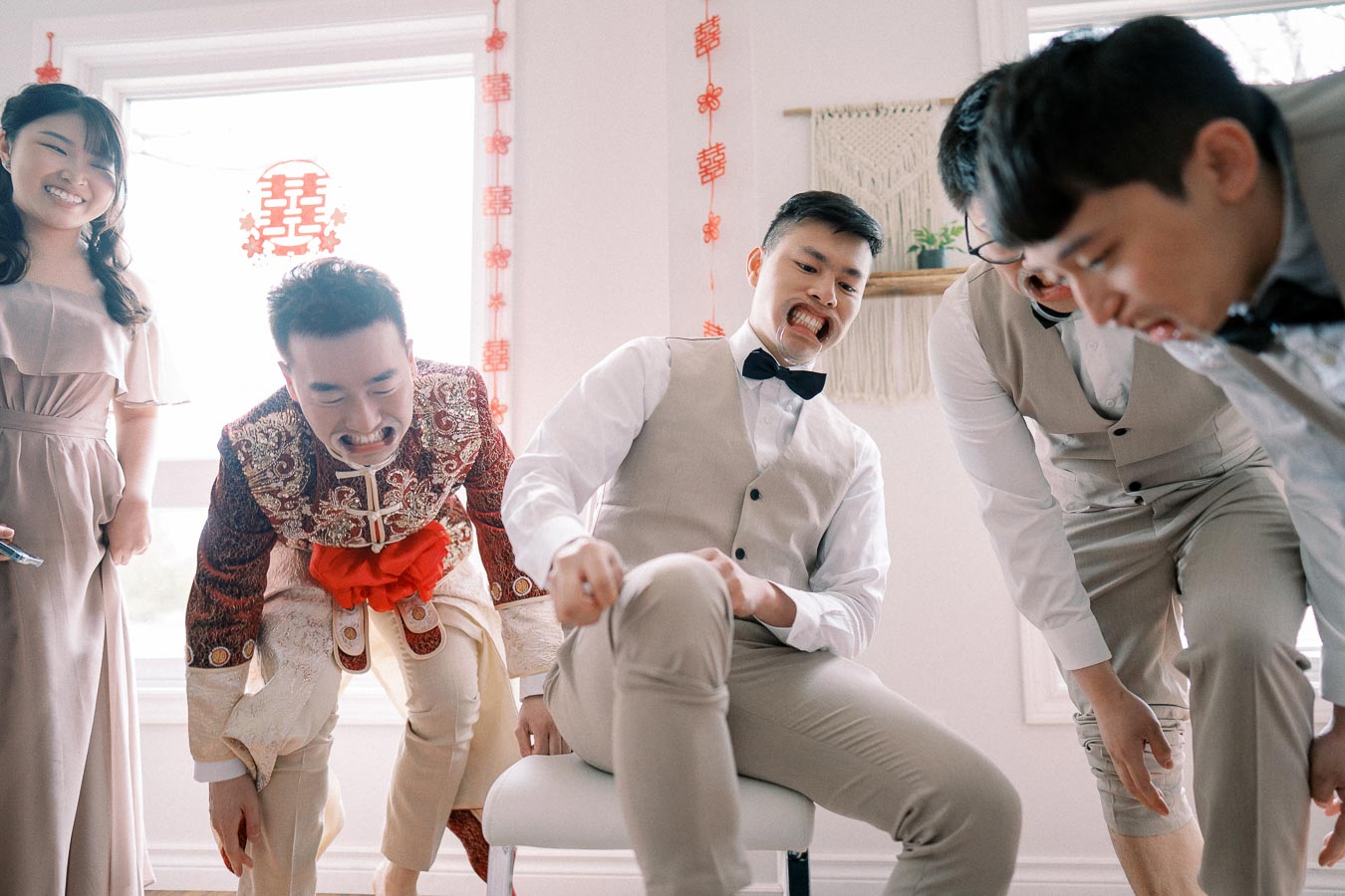 A group of people laughing and leaning forward, wearing formal and traditional attire, in a room decorated with festive red ornaments.