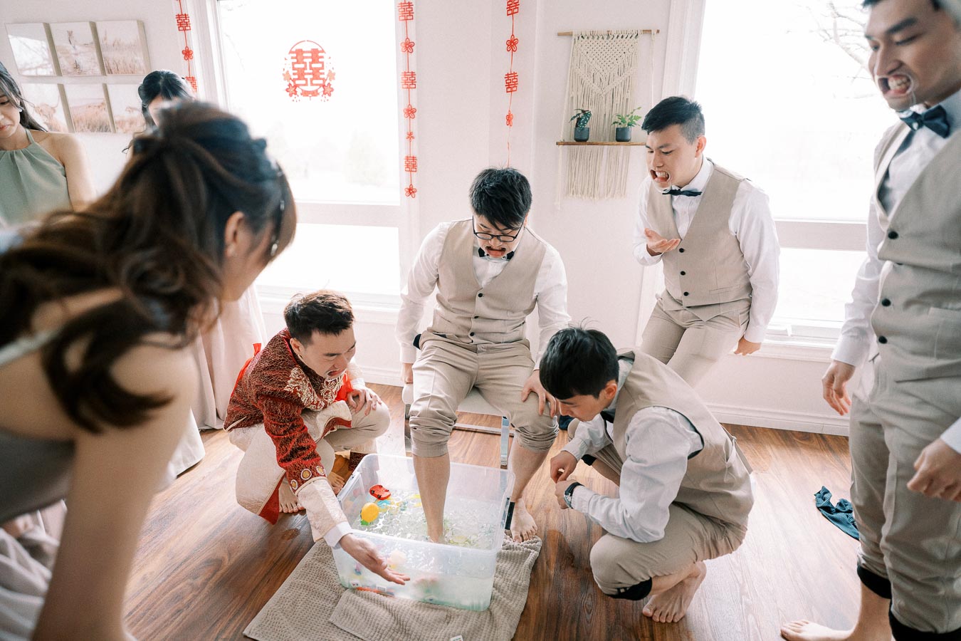 A group of people engaging in a fun wedding or celebration game indoors, with one person dipping their leg into a container filled with water and rubber ducks. Participants are dressed in formal attire, and traditional Chinese decorations hang in the background, creating a lively and festive atmosphere.