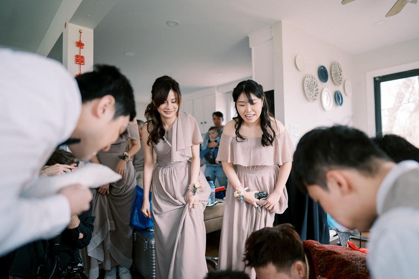 Group of bridesmaids in elegant taupe dresses, smiling indoors during a wedding celebration, surrounded by guests.