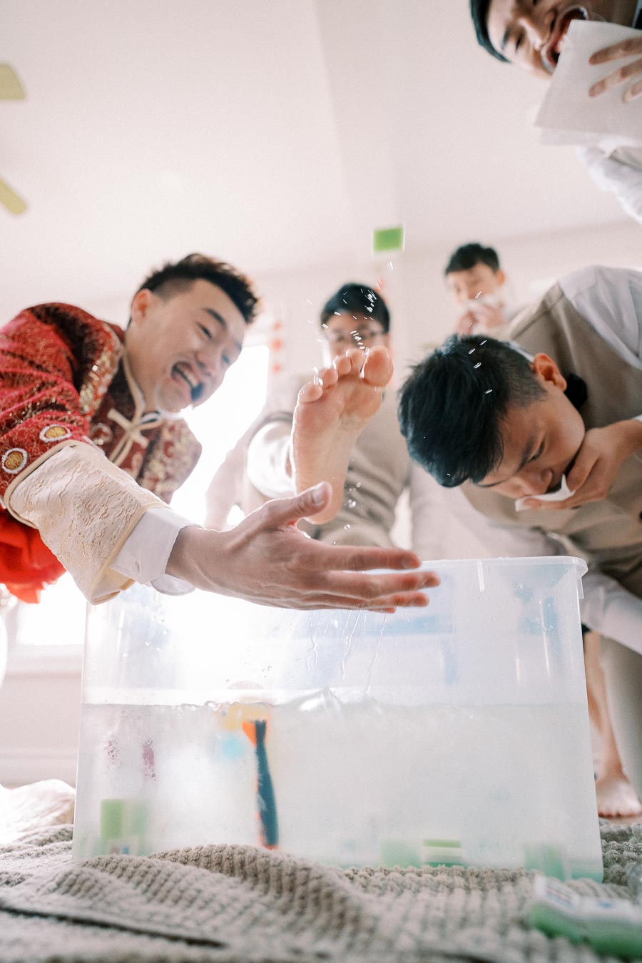 A group of groomsmen in traditional attire participating in a playful wedding game, where one is washing his foot in a plastic tub filled with water, surrounded by others laughing and enjoying the moment indoors.