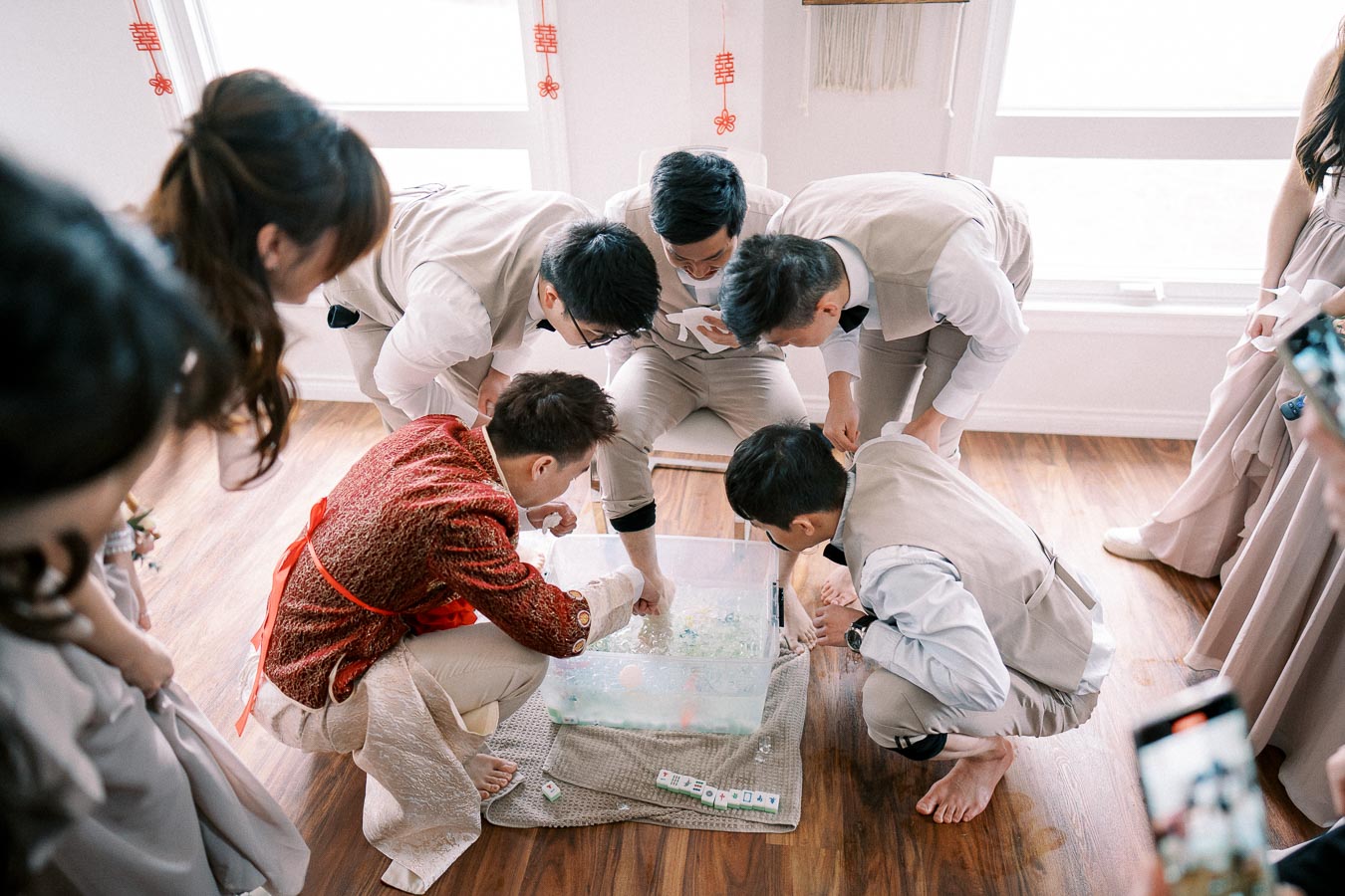 Group of people participating in a traditional game, wearing formal attire, gathered around a plastic container on a wooden floor during a cultural ceremony.