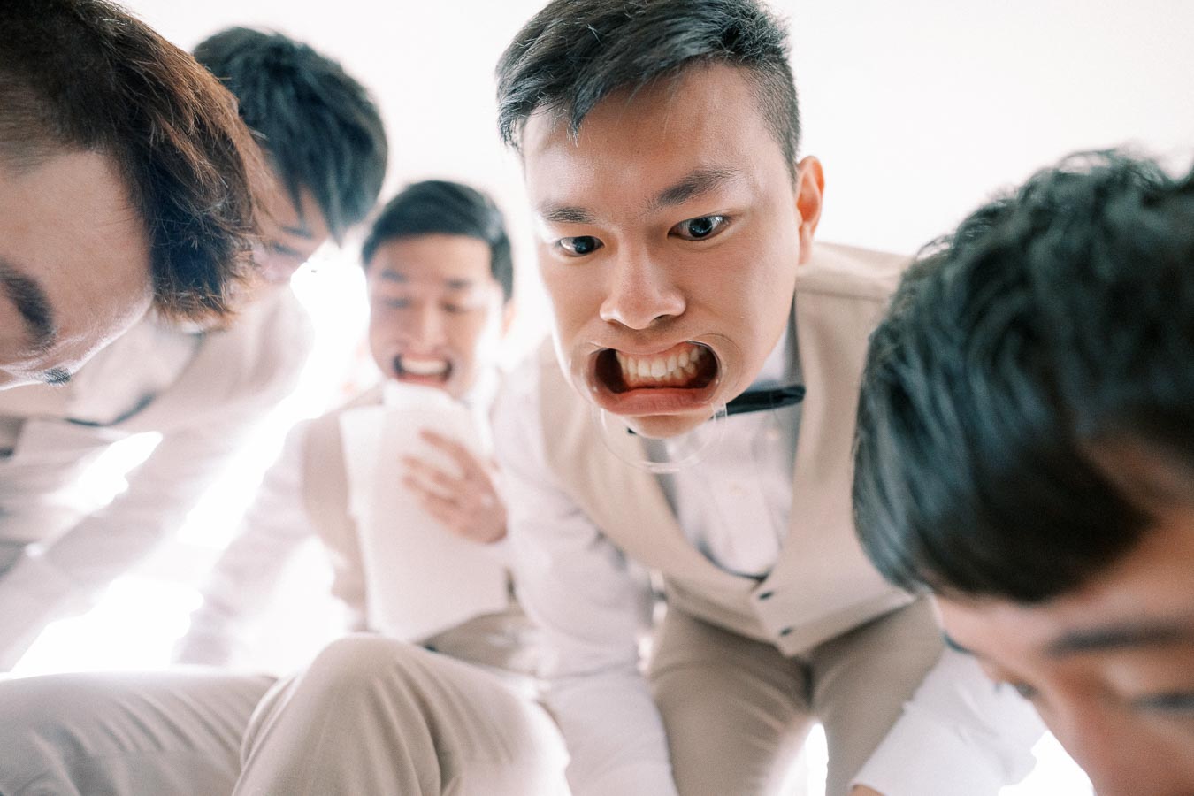Group of laughing groomsmen in suits playing a funny bridal shower game with cheek retractors, creating humorous expressions.