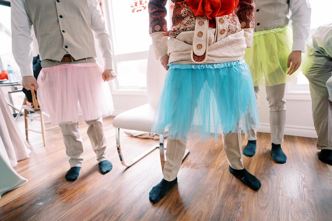 Men having fun and wearing colorful tutus over formal attire at a festive indoor gathering.
