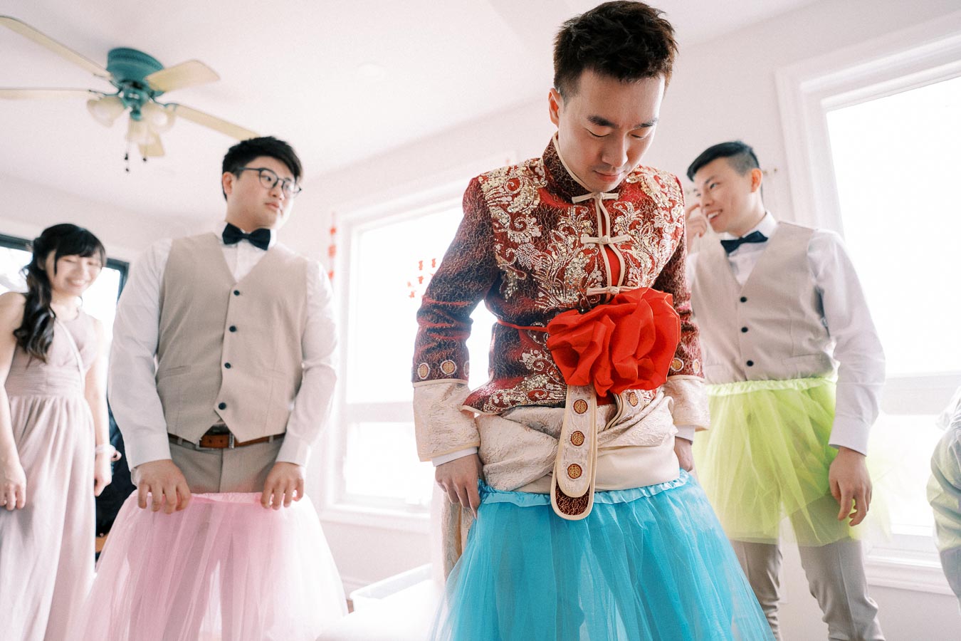 A group of people at a festive event, with a person in traditional ornate attire wearing a playful blue tutu, surrounded by others in formal wear with colorful tutus, highlighting a fun wedding tradition.