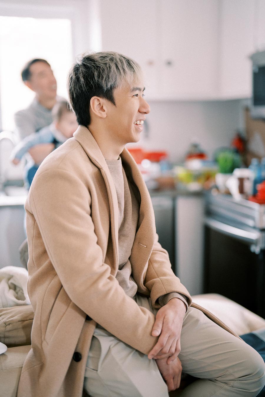 Young man in a beige coat smiling indoors, with blurred family scene in the background involving a parent and child in a kitchen setting.