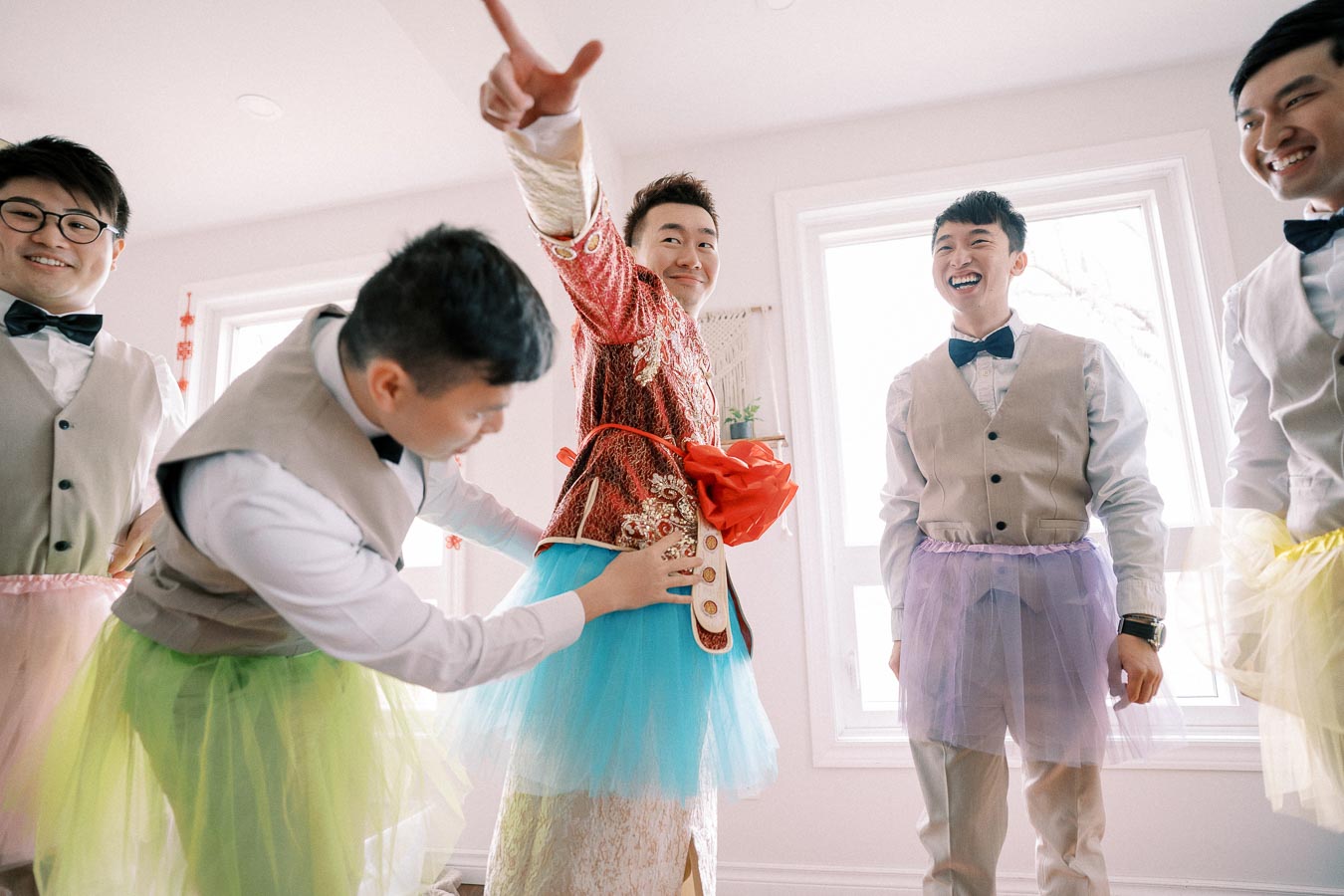 Groom and groomsmen in playful tutus, smiling and enjoying a lighthearted moment before the wedding, in a brightly lit room.