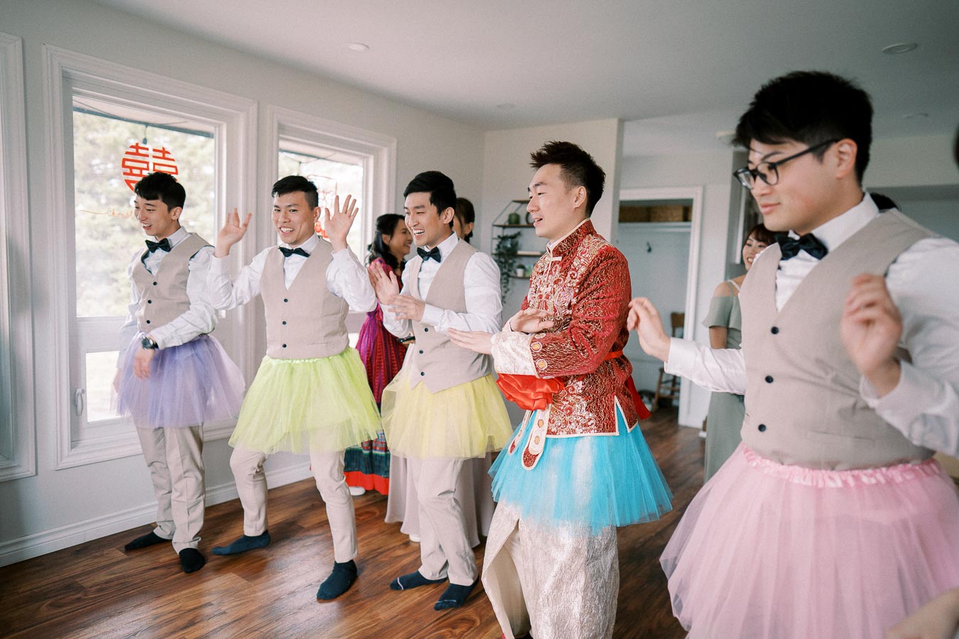 A group of groomsmen wearing colorful tutus and suits, having fun and dancing indoors during a playful wedding tradition.