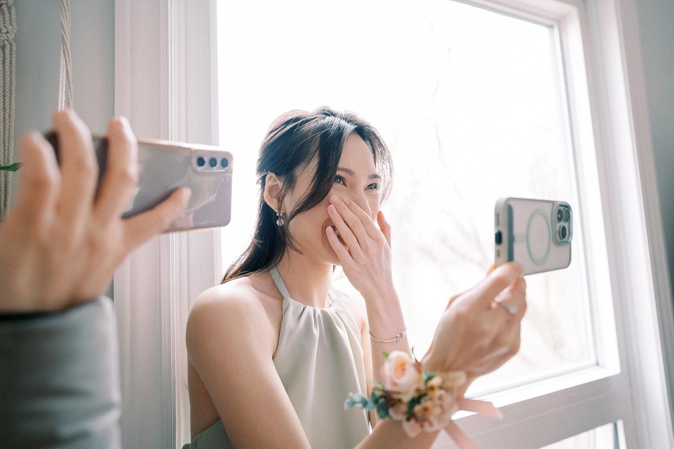 A person wearing a corsage on their wrist is surprised and smiling while looking at a smartphone in front of a bright window, with another phone capturing the moment.