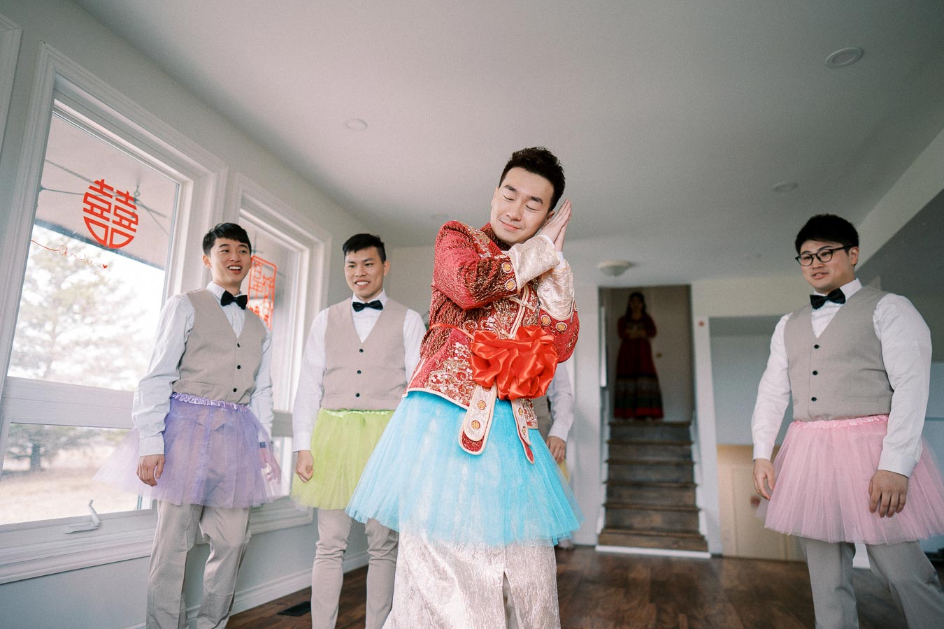 A groom wearing a traditional red robe with a playful blue tutu poses humorously with a sleeping gesture, while three groomsmen in vests and colorful tutus smile and watch, creating a fun and lighthearted wedding moment indoors.