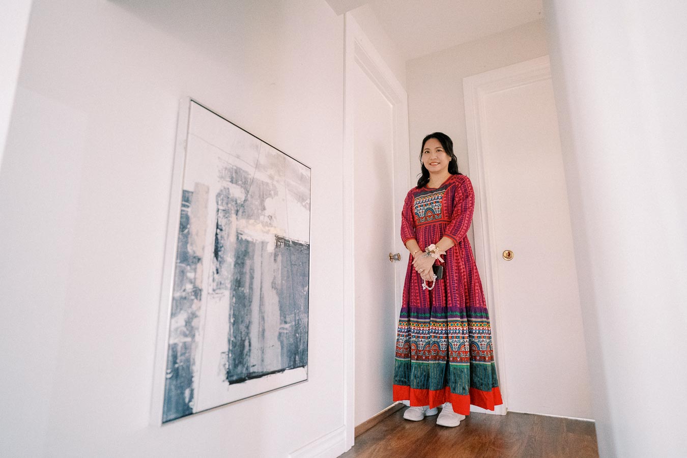 A woman in a colorful traditional dress stands in a brightly lit hallway next to an abstract painting on the wall.