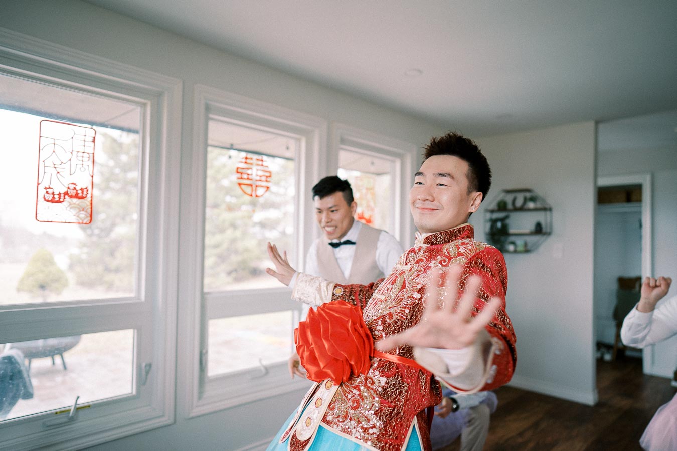 A person in traditional Chinese wedding attire playfully posing indoors, with decorative red Chinese symbols on the windows, celebrating a joyful occasion.