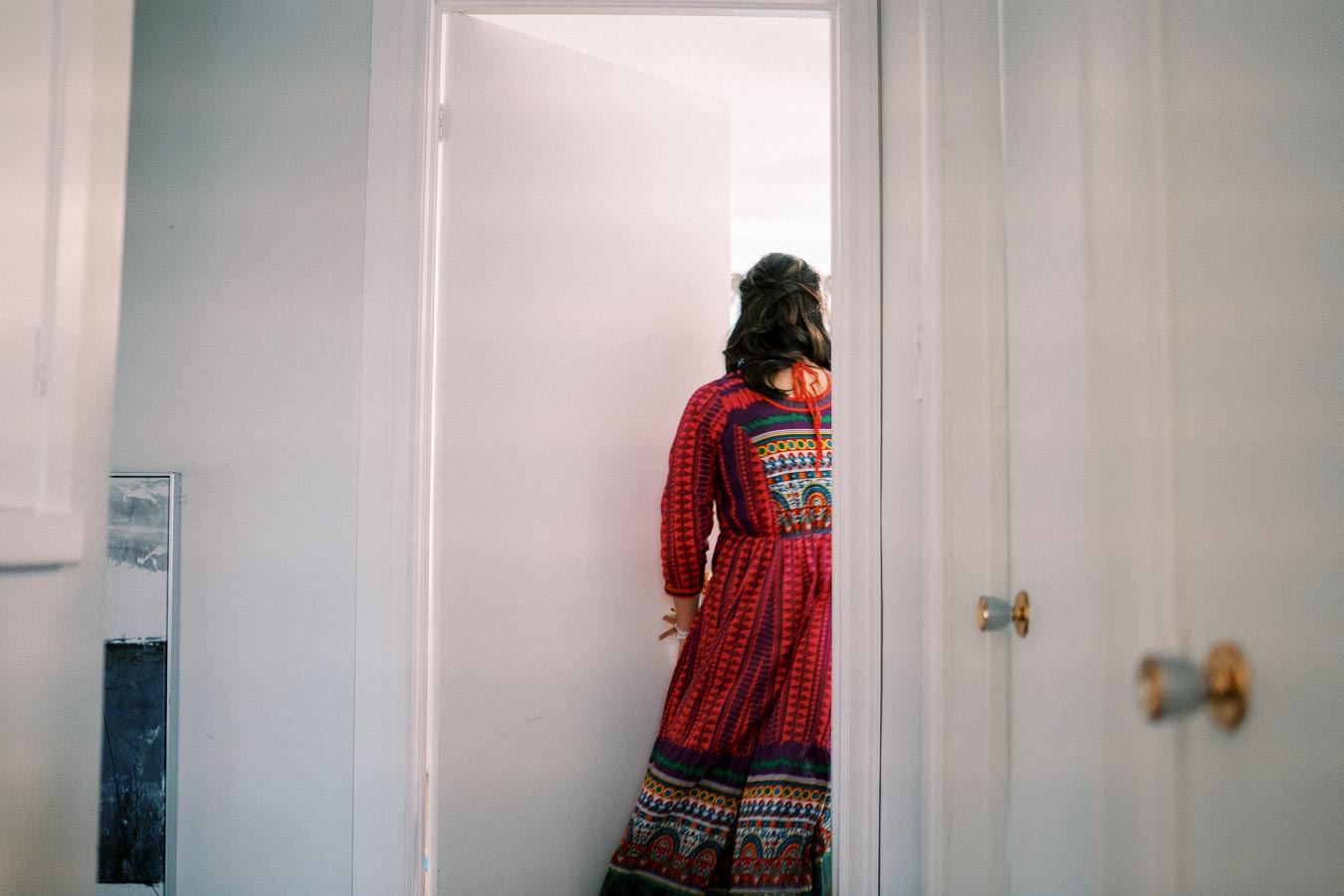 A woman in a vibrant, patterned red dress stands partially seen through an open door in a softly lit room.