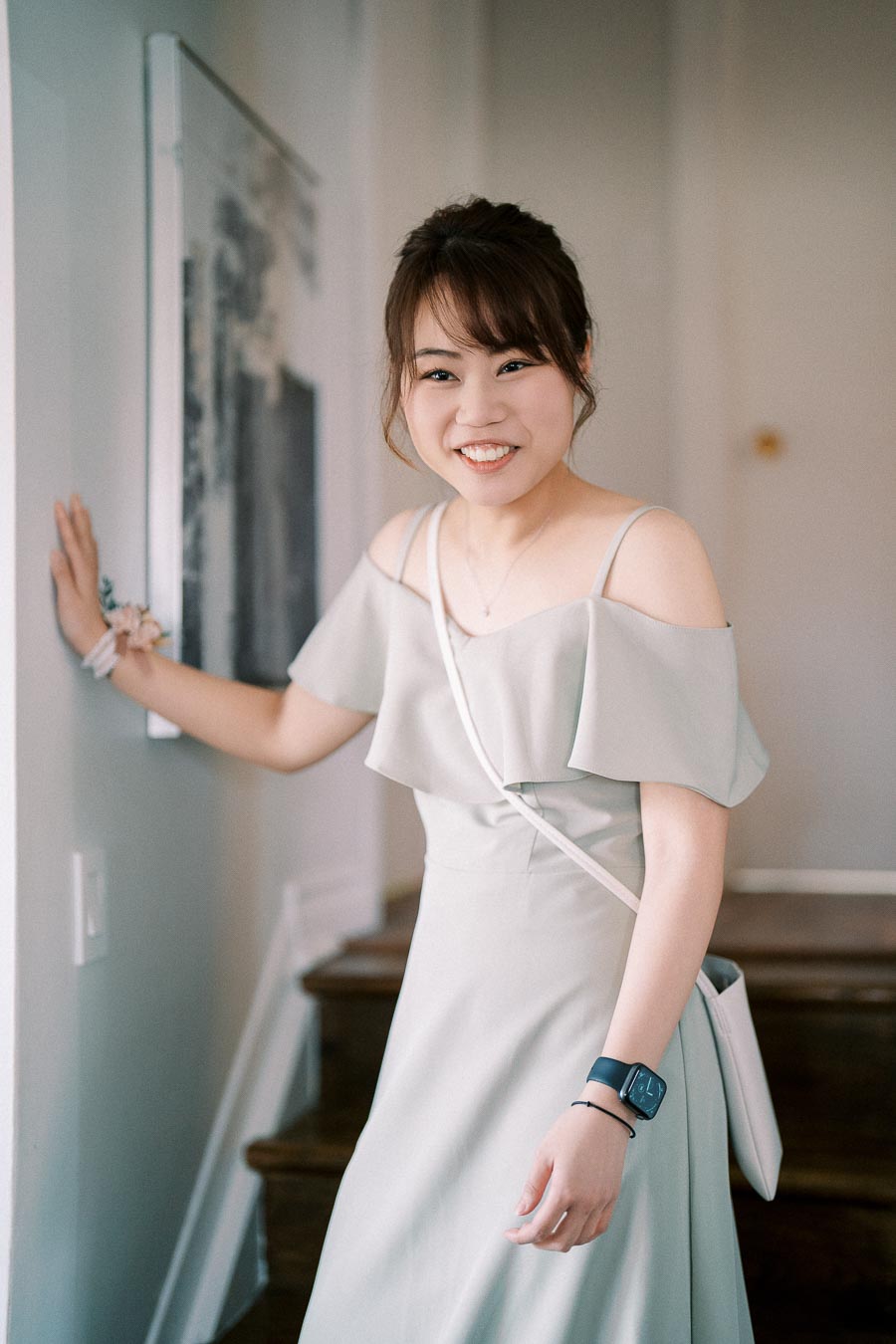 Young woman smiling in a light gray off-shoulder dress, standing indoors near a staircase, wearing a smartwatch and a small white crossbody bag.