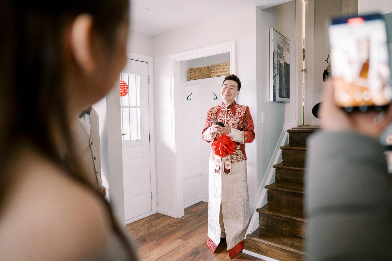 A person smiling and wearing traditional Chinese wedding attire, standing in a modern hallway, with people capturing the moment on their phones.