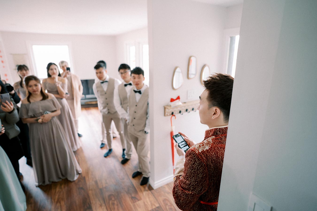 A group of elegantly dressed people, including women in gowns and men in suits, gather in a well-lit room with hardwood flooring, while a man in a red traditional outfit stands to the side holding a phone.