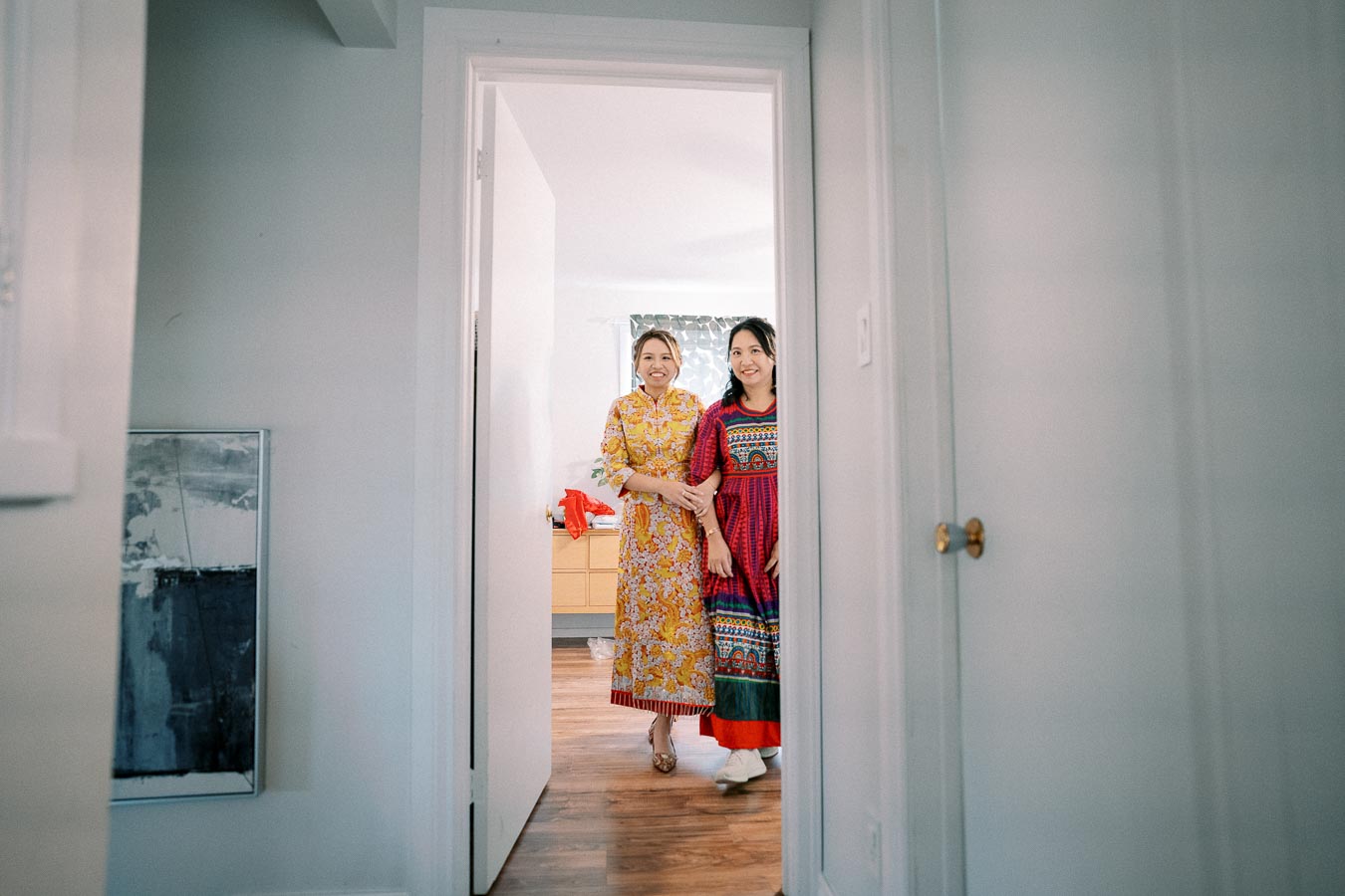 Two women in colorful traditional dresses standing in a doorway, smiling warmly at the camera, inside a bright and cozy room.