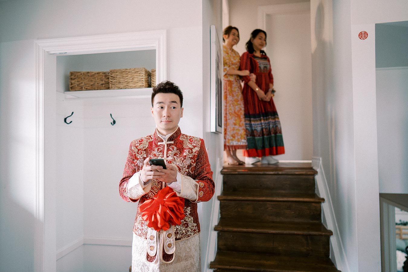Young man in traditional Chinese wedding attire with red embroidery, holding a phone, standing near stairs with two women in colorful dresses observing from above.