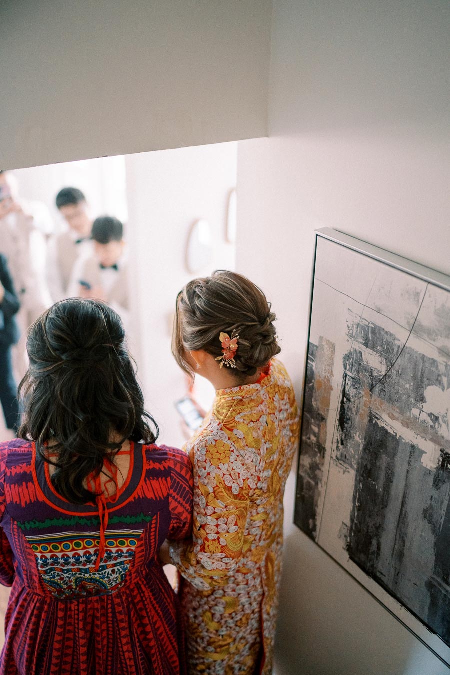Two women seen from behind, wearing colorful traditional attire with intricate patterns, standing indoors by a modern abstract painting.
