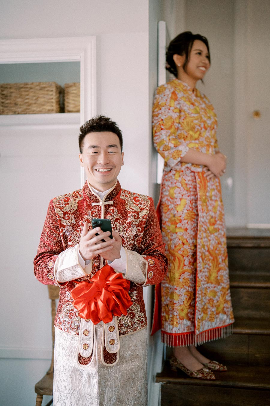A man in traditional, ornate red and gold attire holds a phone and smiles, while a woman in a colorful dress stands in the background, creating a joyful and festive atmosphere.