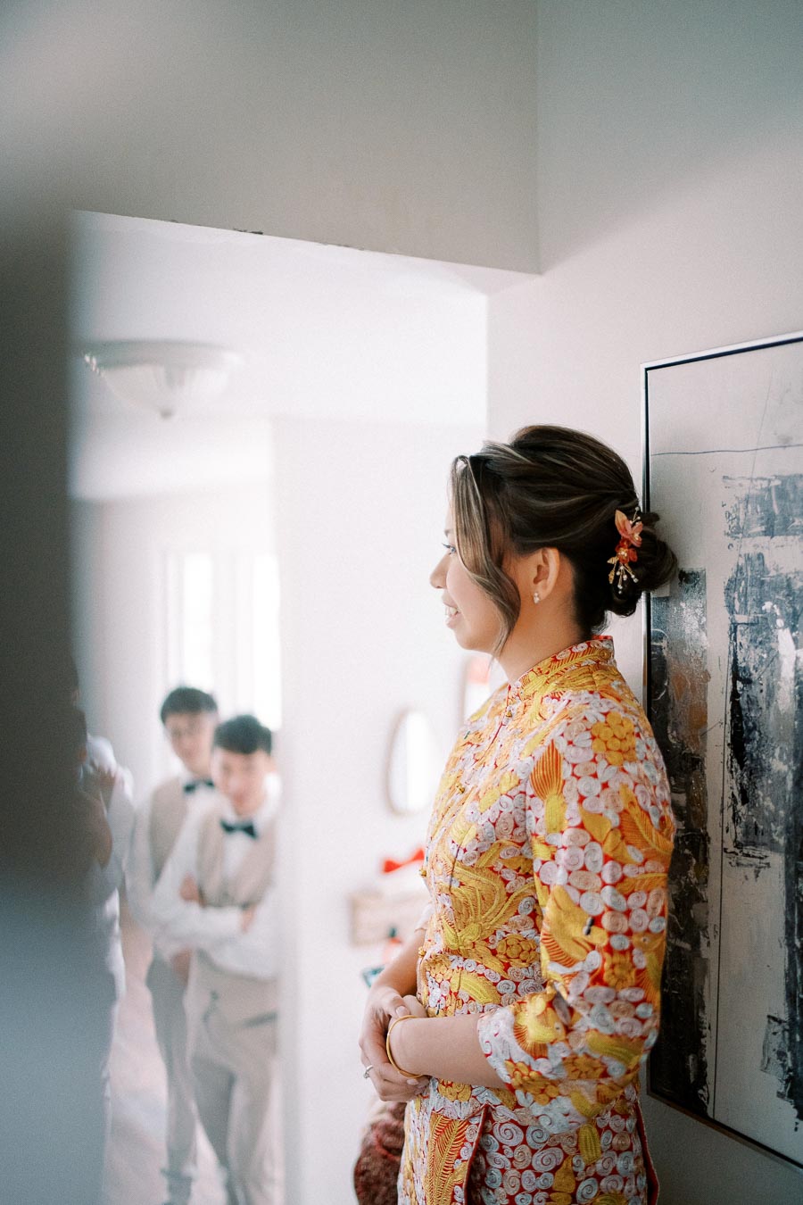 A woman in a vibrant yellow and red traditional dress stands smiling in a modern home, with a blurred background showing three people dressed in formal attire.